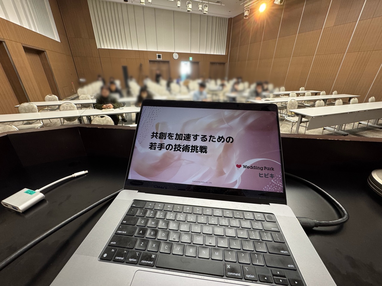 登壇台から会場を見渡した様子。MacBook Proの画面に登壇スライドの表紙の「共創を加速するための若手の技術挑戦」が表示されている。